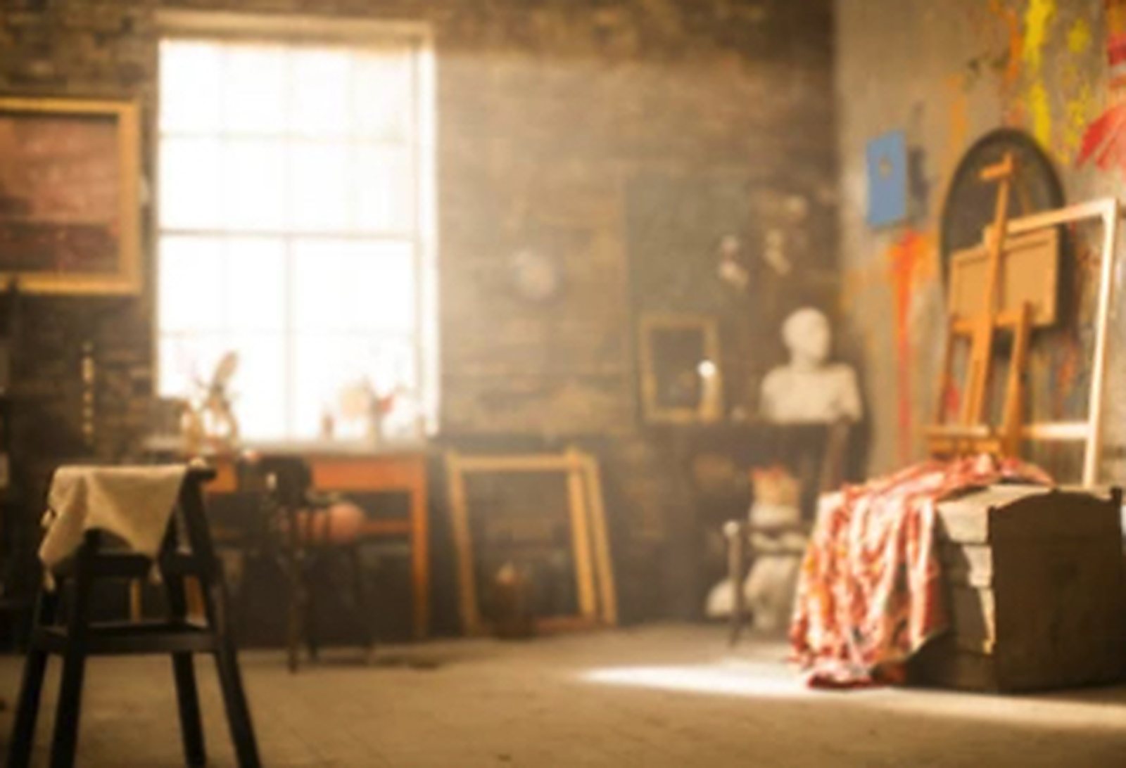 A working artist's studio with an easel, stacked canvases and daylight from a tall window.