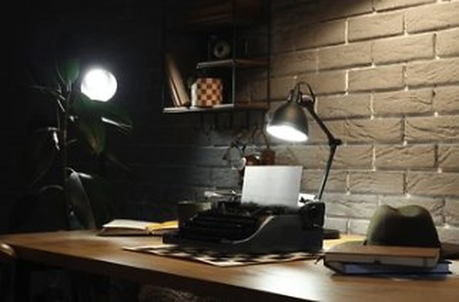 A typewriter, notebooks and a desk lamp on a wooden writing desk against a brick wall.