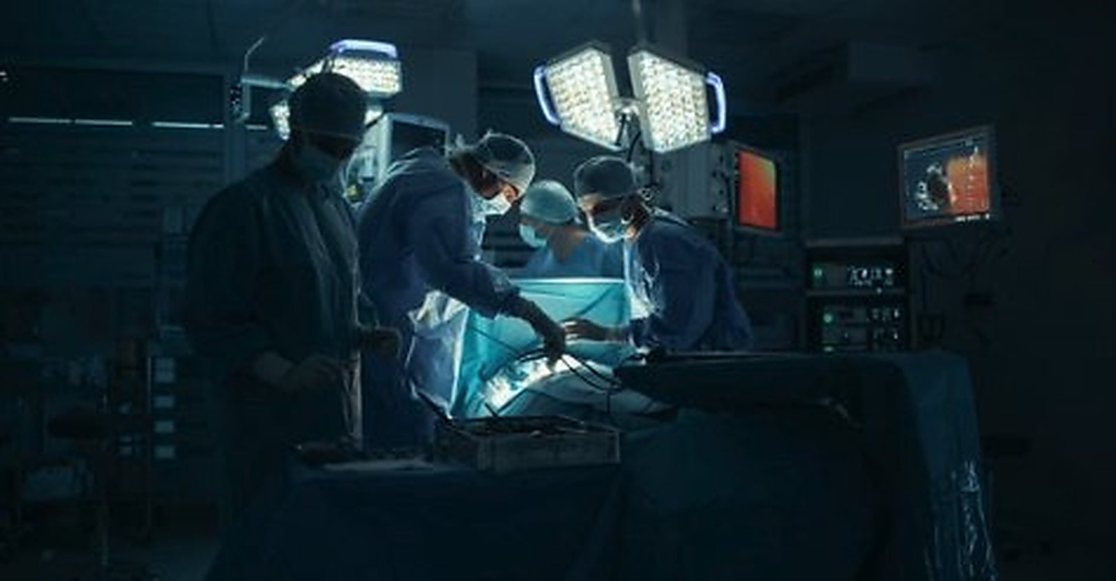 A gowned surgical team working under theatre lamps in a darkened operating room.