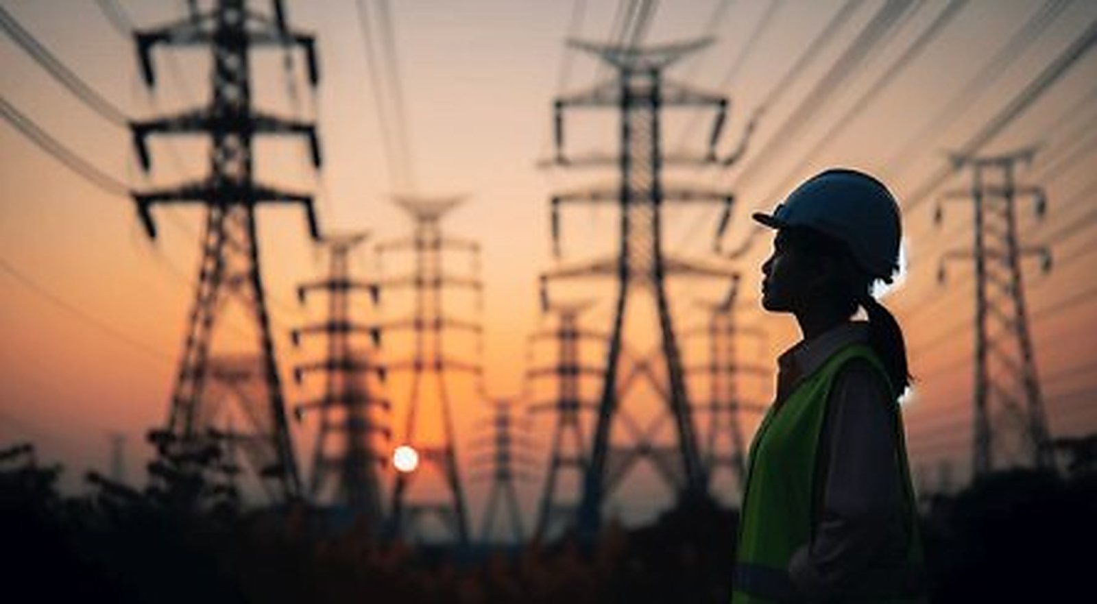 An engineer in a hard hat and high-visibility vest silhouetted against transmission pylons at sunset.