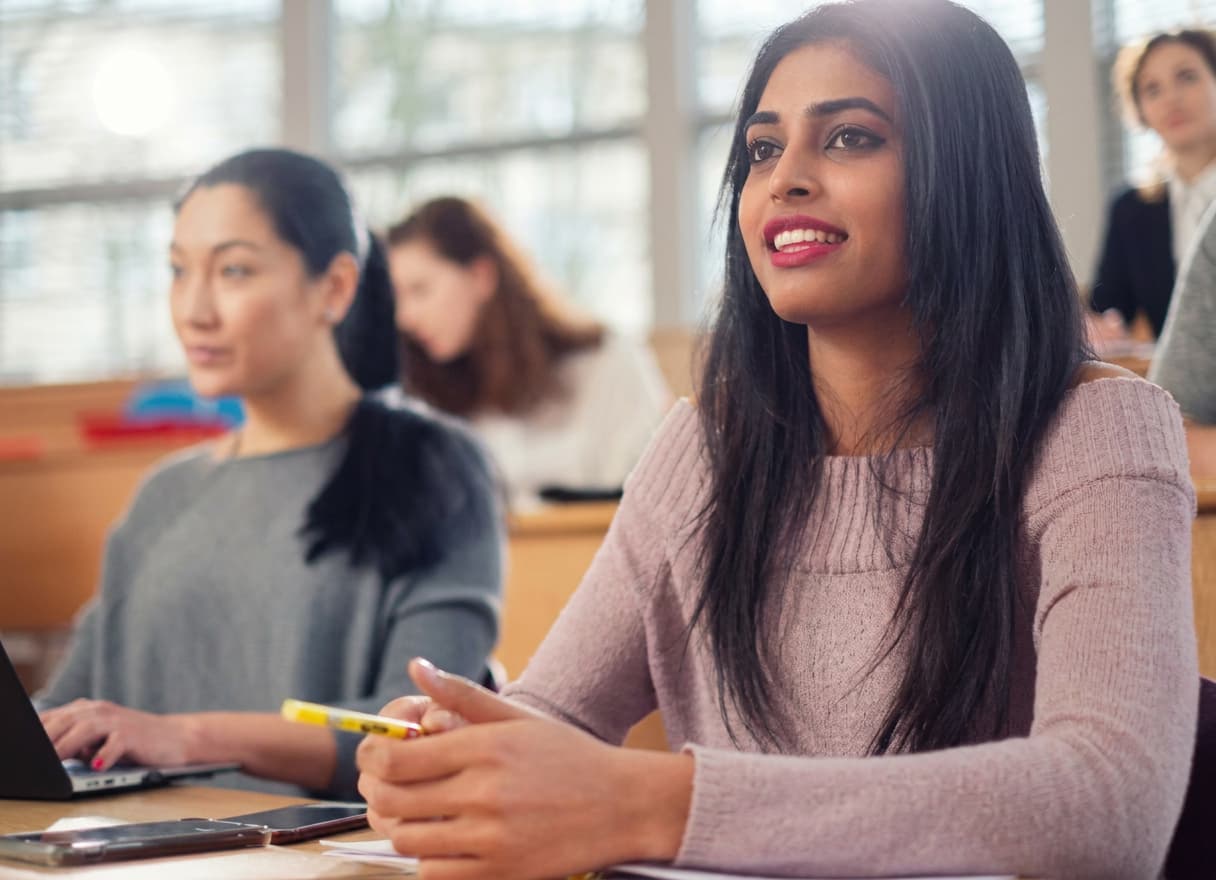 A student listening intently in a bright classroom, pen in hand, classmates working behind her.
