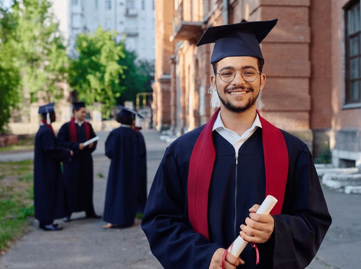 A smiling graduate in a navy gown and crimson stole holding a rolled diploma.