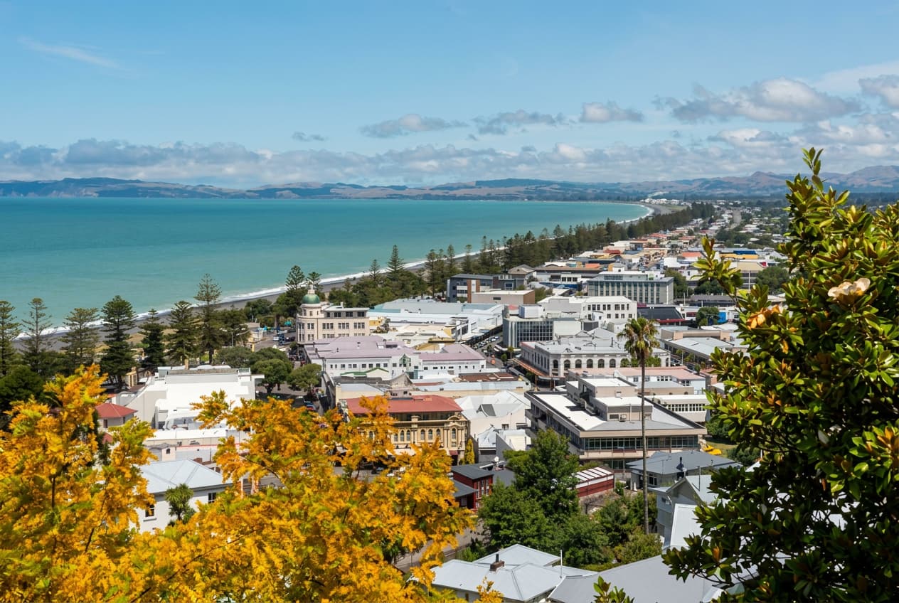 A coastal New Zealand town seen from above with a sweeping teal bay and distant hills.