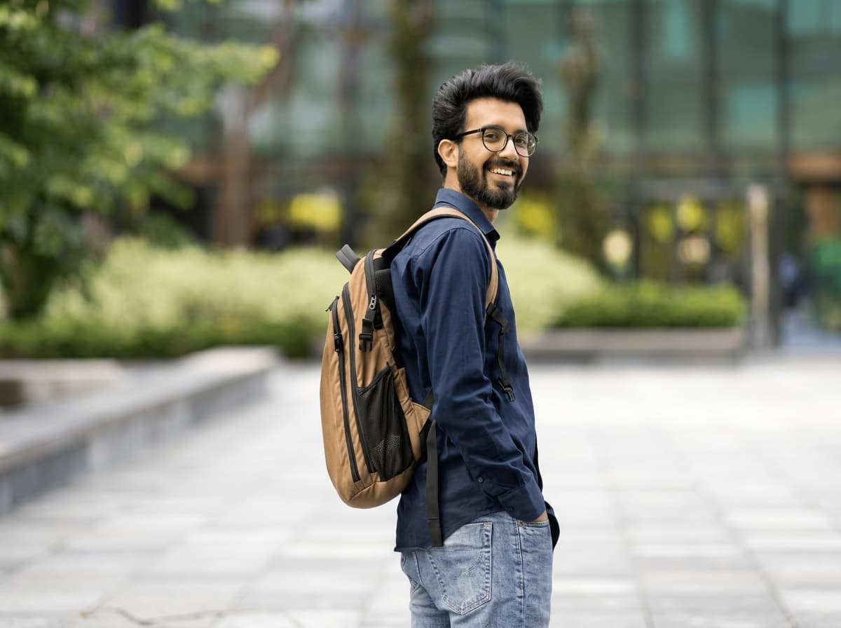 A student with a tan backpack turning to smile over his shoulder on a paved campus plaza.
