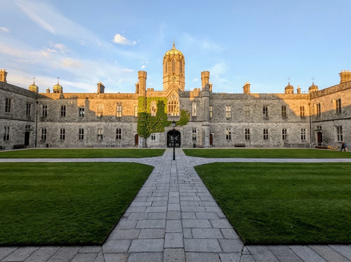 A grey limestone university quadrangle with a green copper clock tower and wide lawns in evening sunlight.