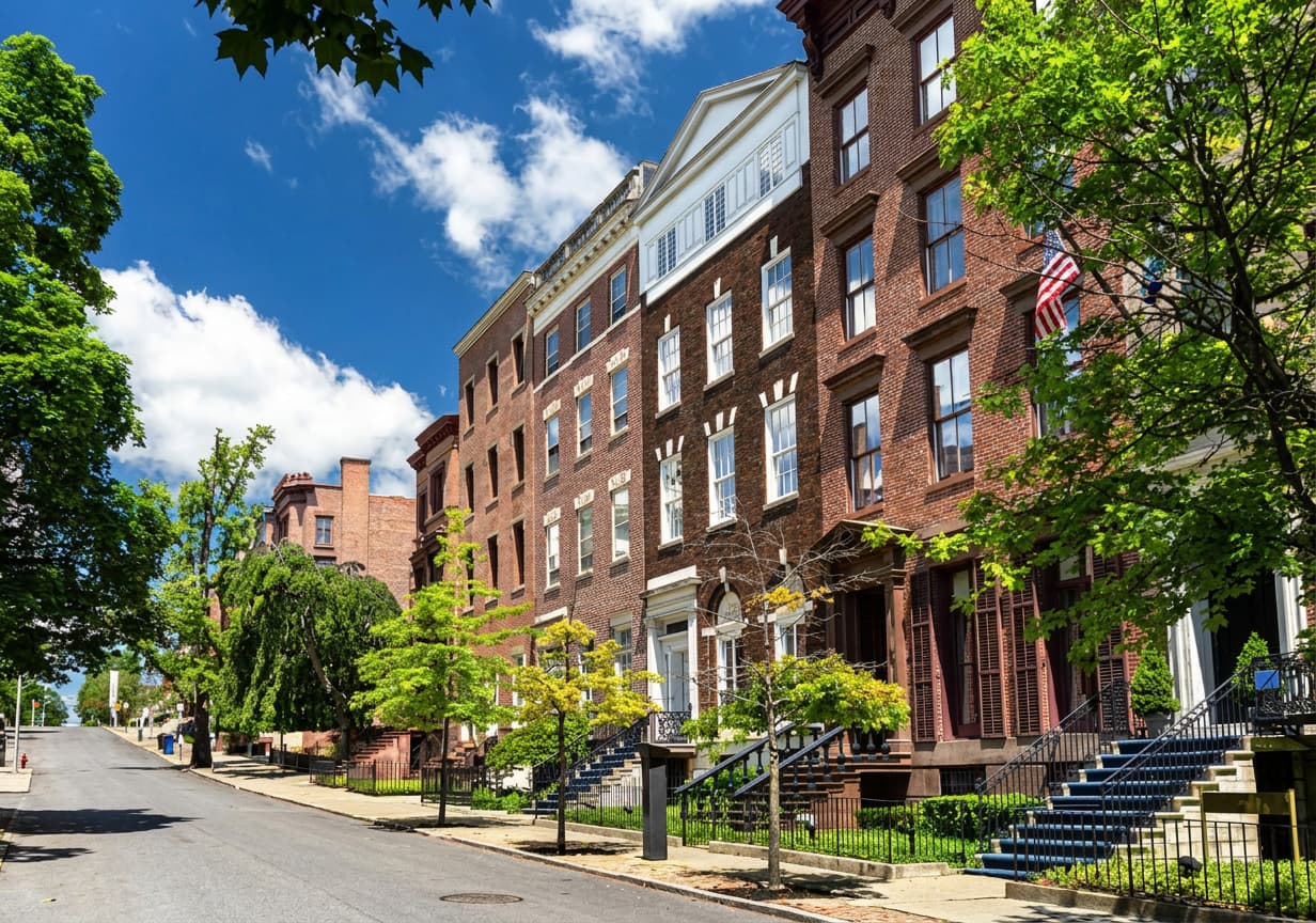 A sunlit street of red-brick American townhouses with stoops and an American flag.