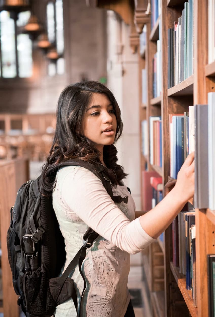 A student reaching for a book on tall library shelves.