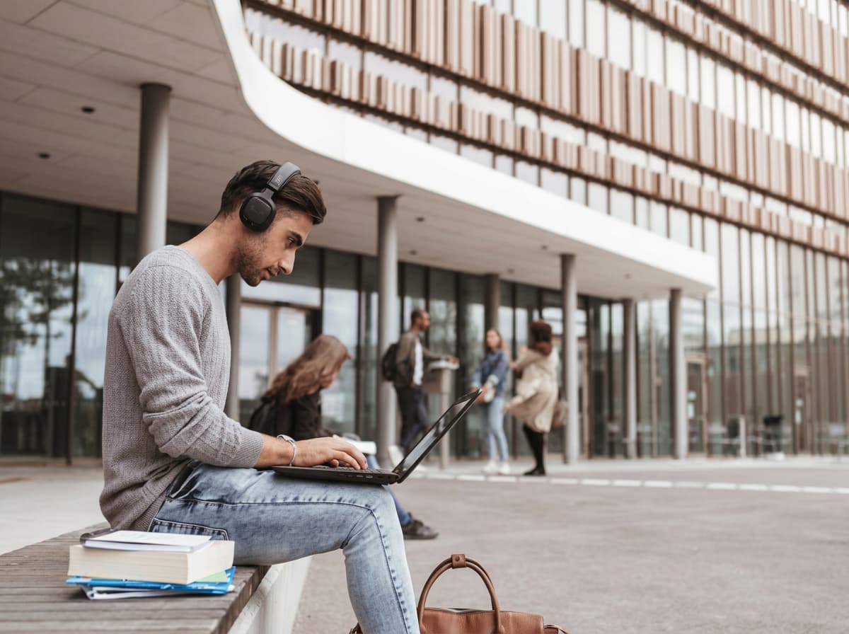 A student in headphones working on a laptop outside a modern building.