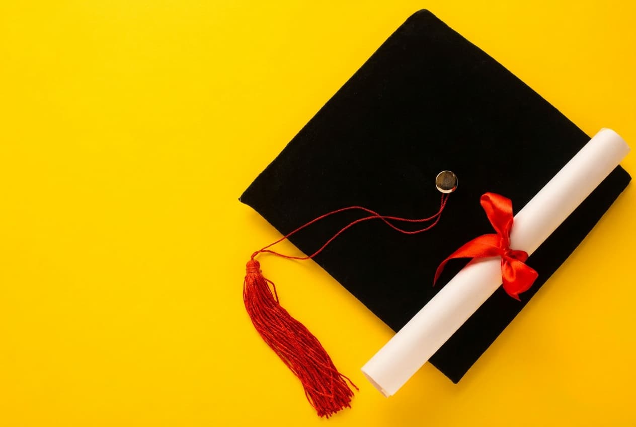 A graduation cap and ribbon-tied diploma arranged on a bright yellow background.