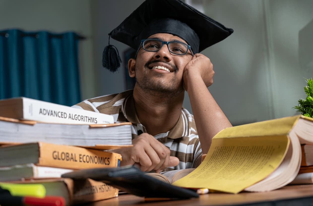 A student in a graduation cap beside advanced textbooks.