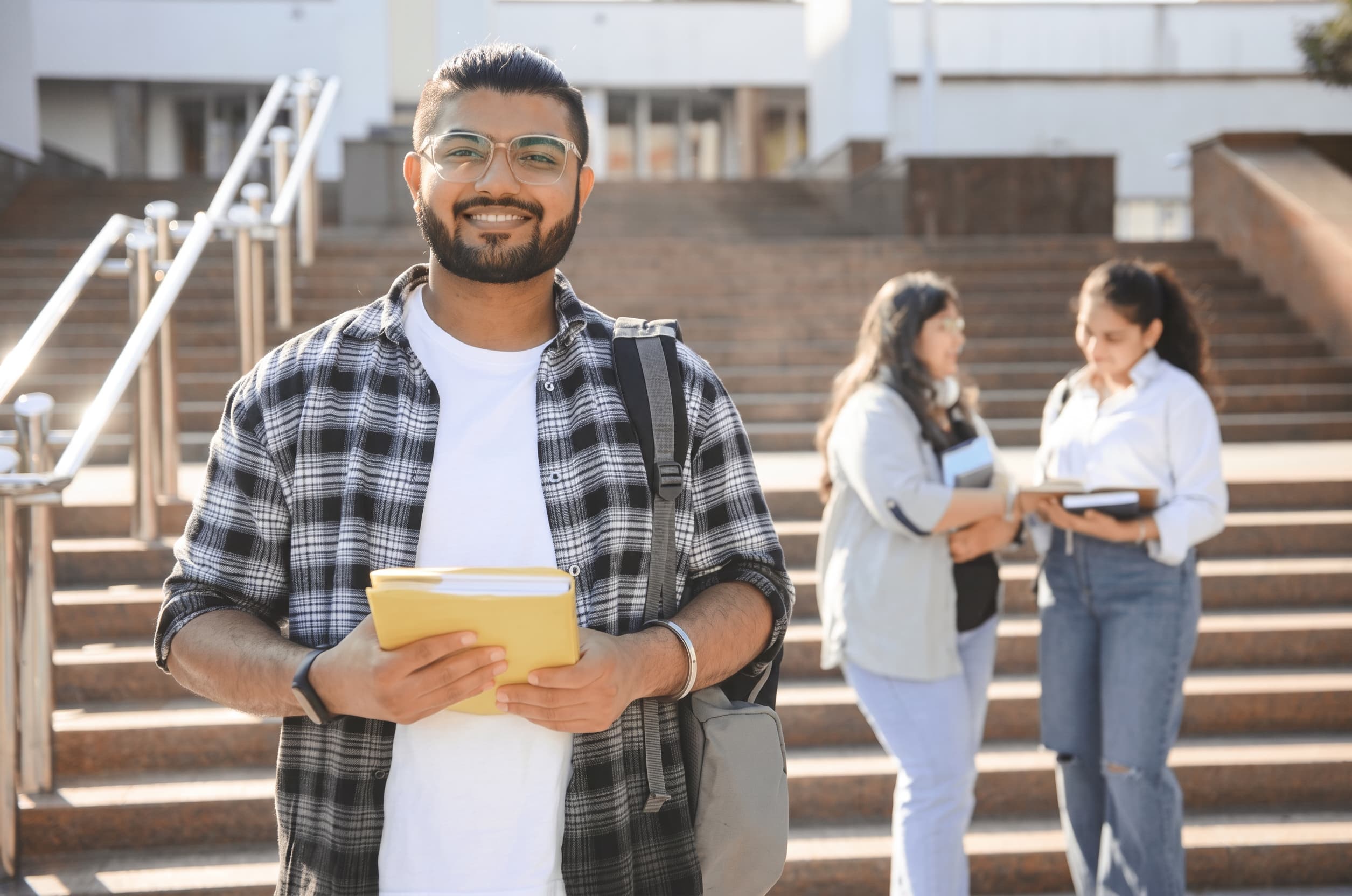 A student standing on wide campus steps in bright sunshine, holding a folder.