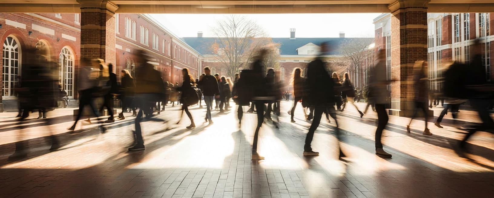 Students silhouetted in a backlit brick colonnade opening onto a campus quad.