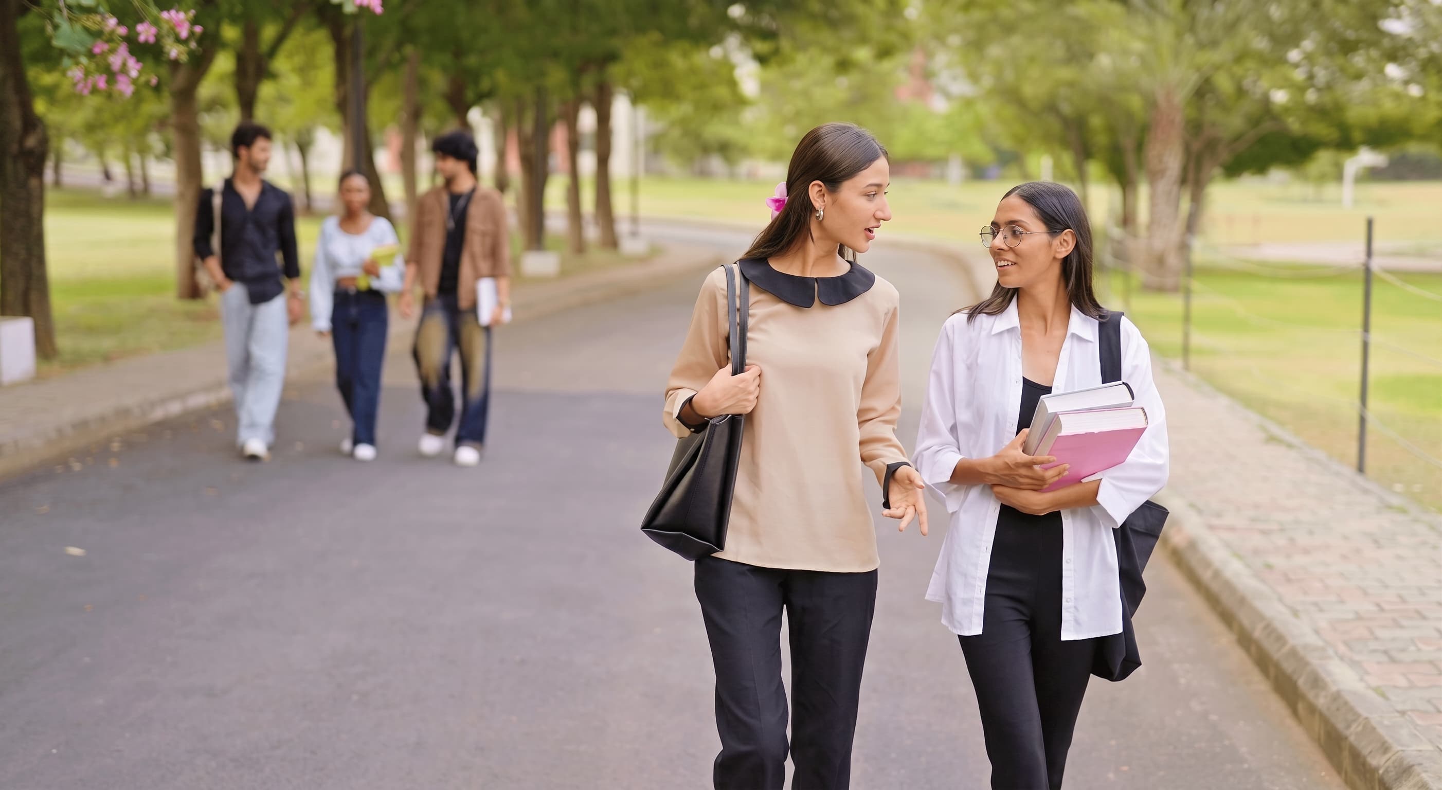 Two students walking and talking along a tree-lined university avenue in summer.