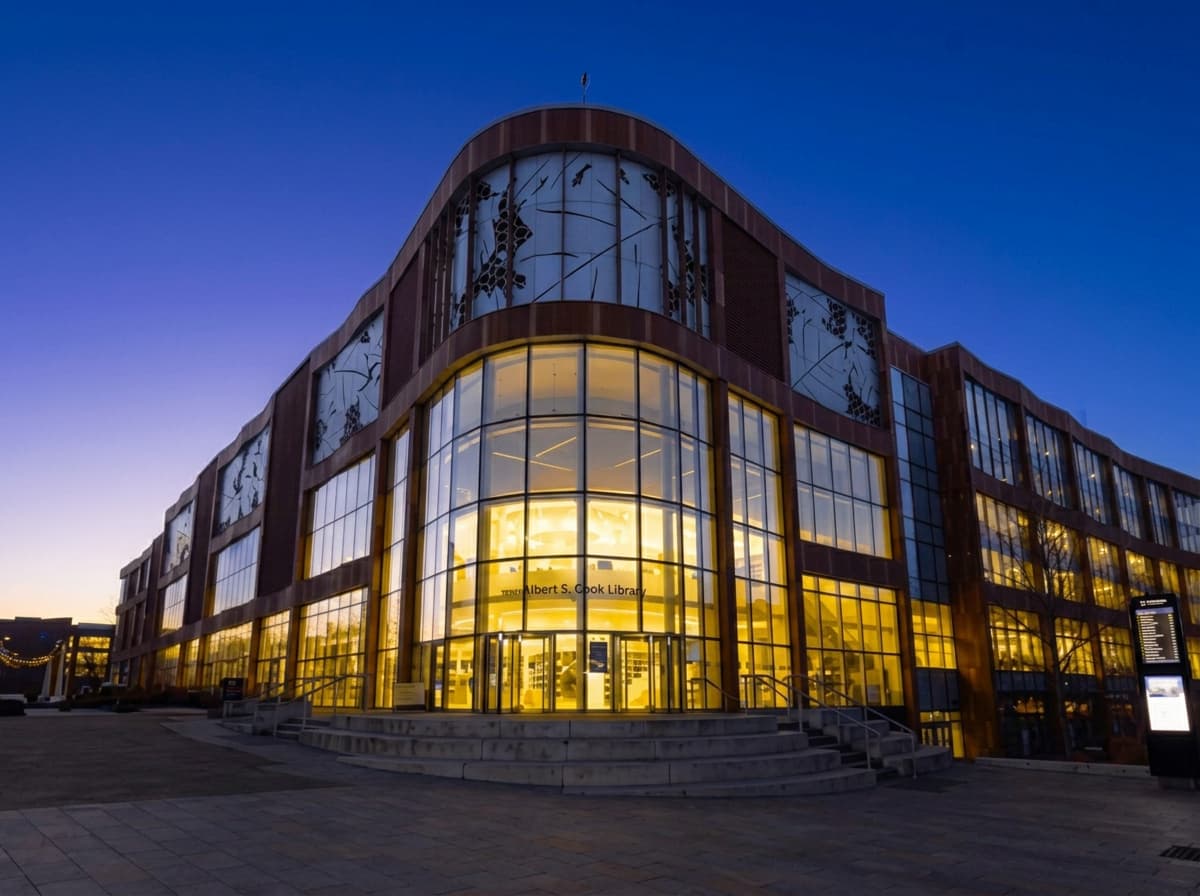A curved glass university library glowing gold against a deep blue evening sky.